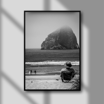 A black and white photograph in a frame, depicting a man sitting on a beach looking towards the horizon, with a large rock formation in the distance.